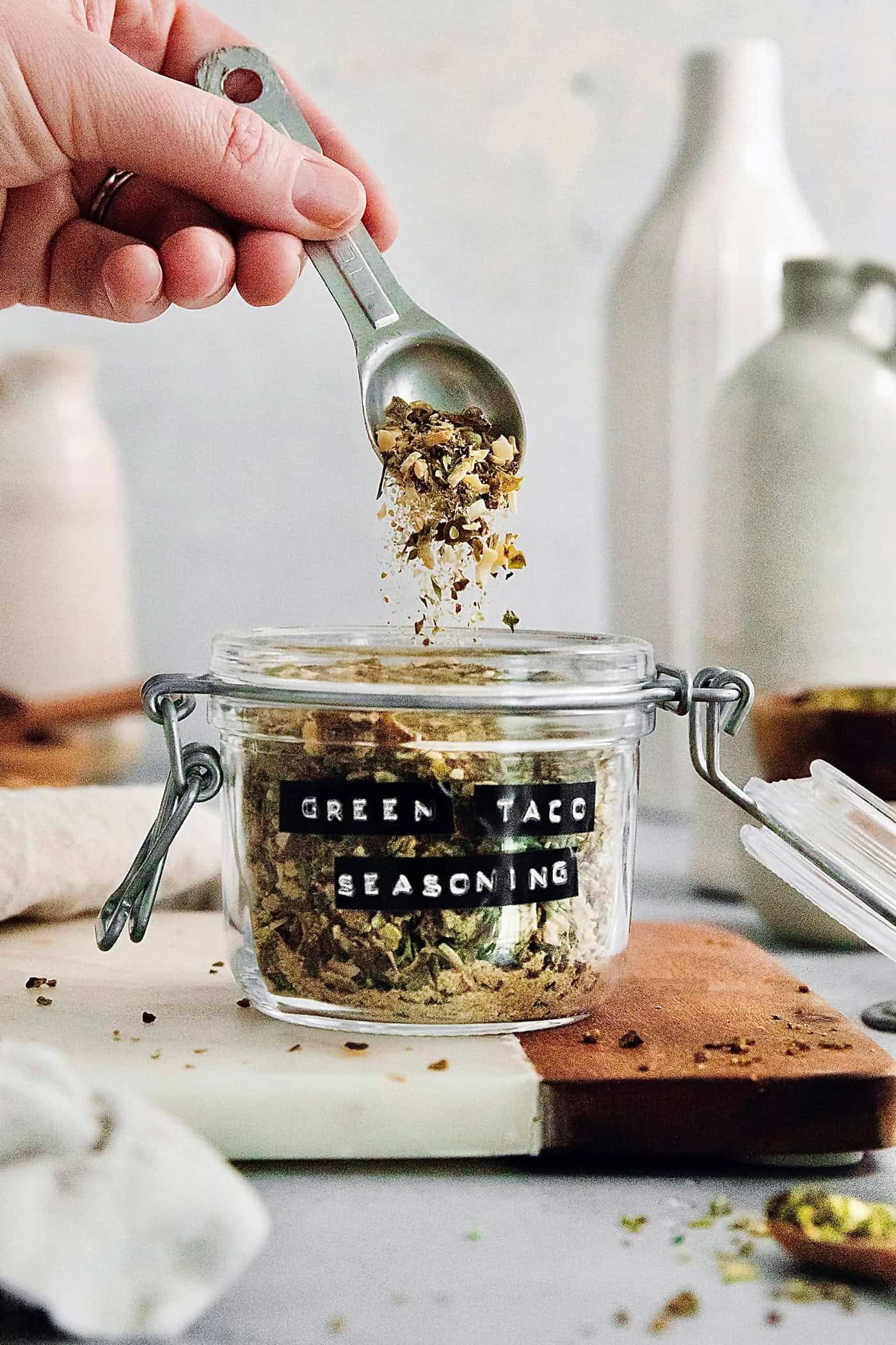 a hand holding a spoon that is dropping green taco seasoning into a jar