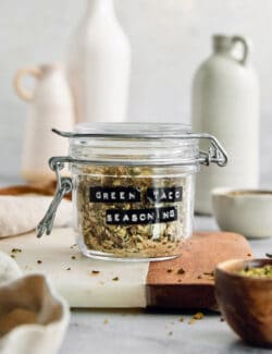 A labeled glass jar of green taco seasoning on a cutting board.