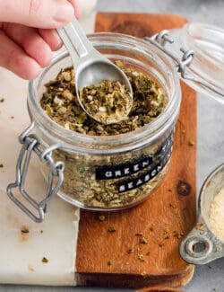 A hand dip a spoon into a jar of green taco seasoning.