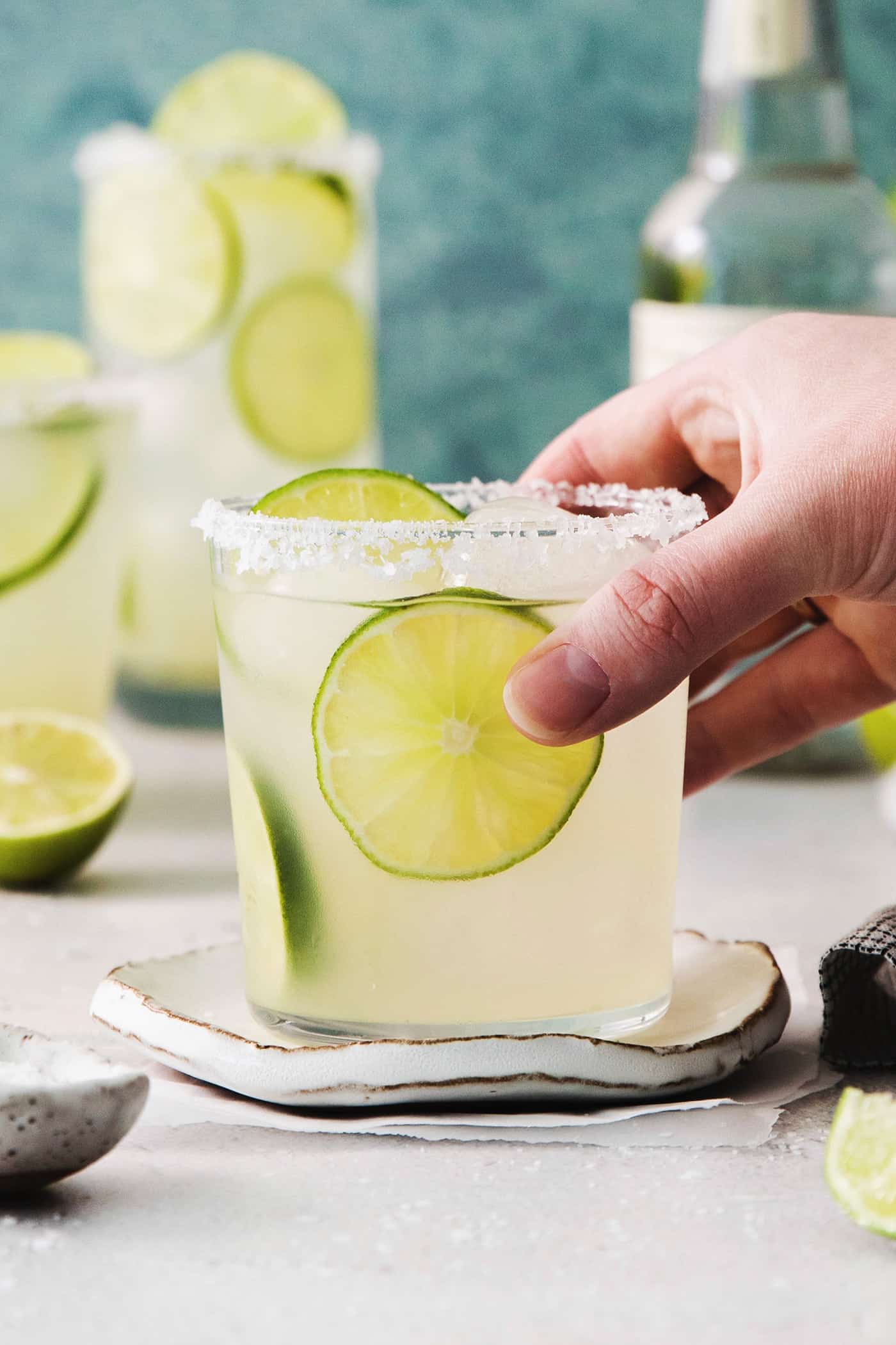 A hand reaches to pick up a glass of classic margarita with more margaritas in the background.