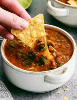 A hand dips a tortilla chip into a bowl of warm salsa.