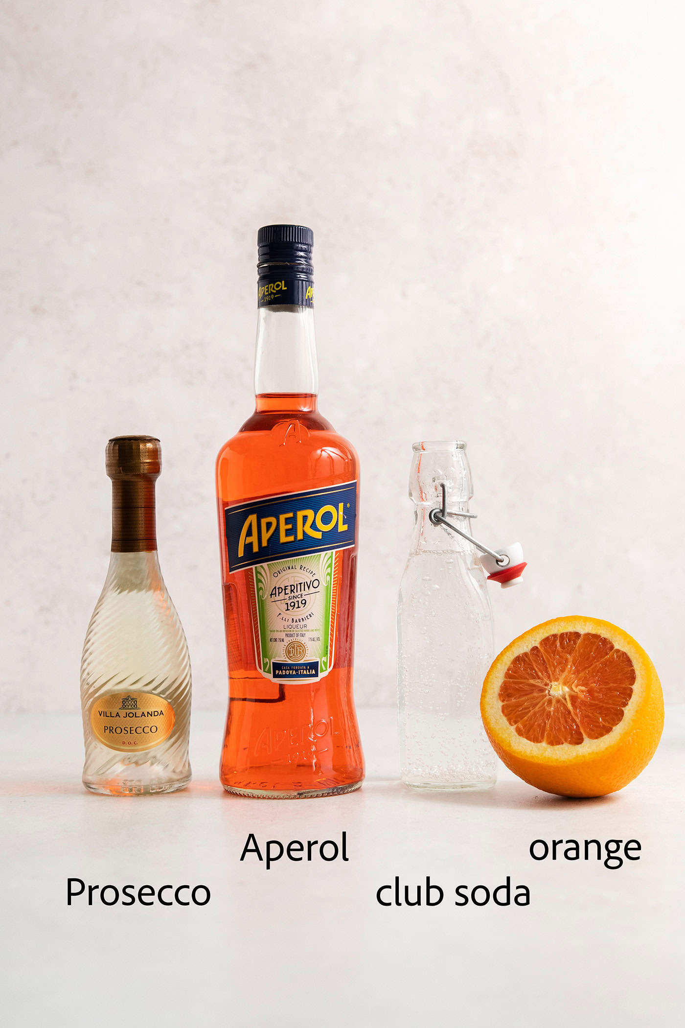 Aperol spritz ingredients are shown and labelled: a bottle of prosecco, a bottle of Aperol, club soda, an orange.