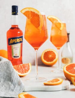 Glasses of Aperol spritz are surrounded with orange wedges next to a bottle of Aperol.