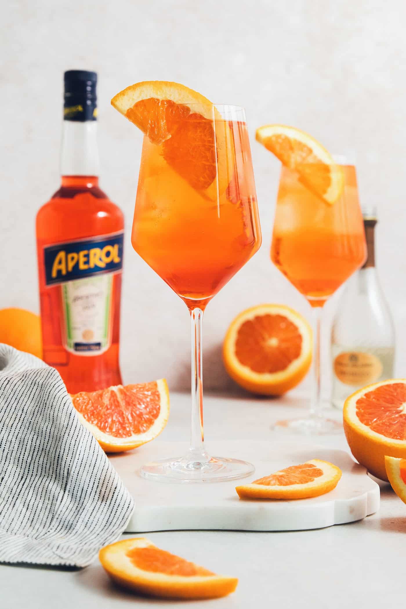 Glasses of Aperol spritz are surrounded with orange wedges next to a bottle of Aperol.