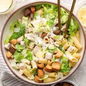 A big bowl of Caesar chicken pasta salad with serving utensils.