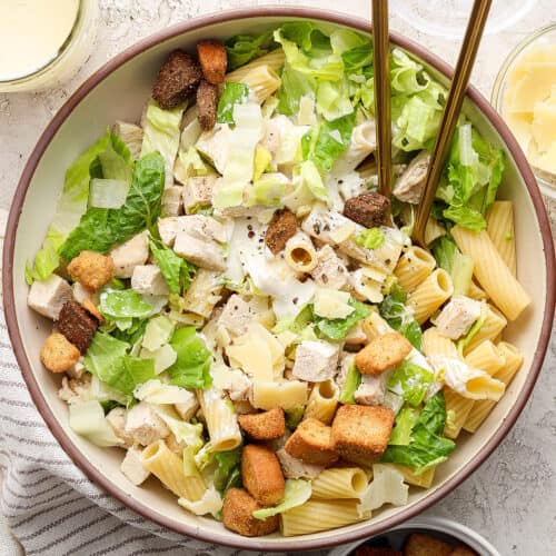 A big bowl of Caesar chicken pasta salad with serving utensils.