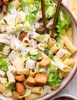 A side view of a bowl of Caesar chicken pasta salad with serving spoons.