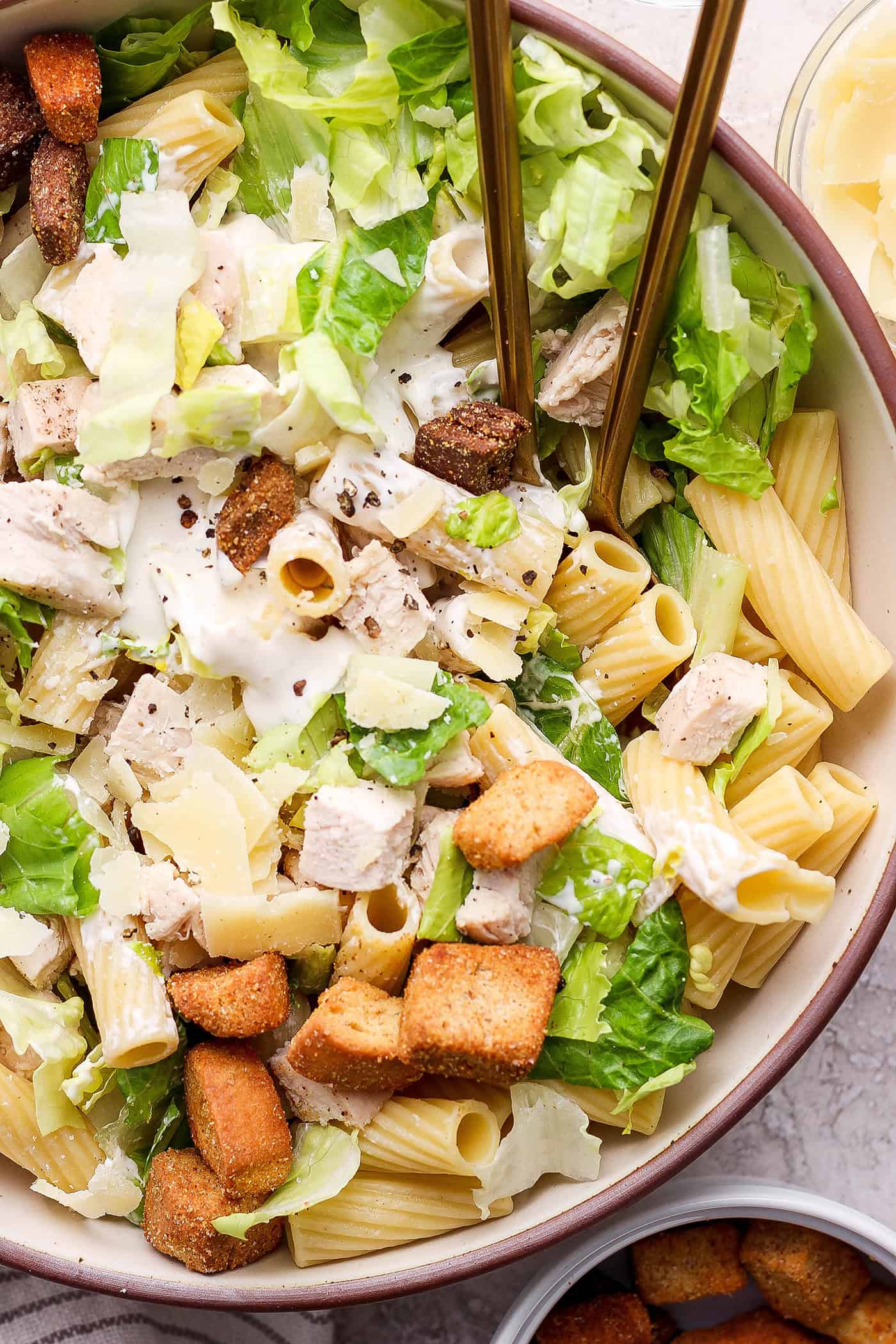 A side view of a bowl of Caesar chicken pasta salad with serving spoons.