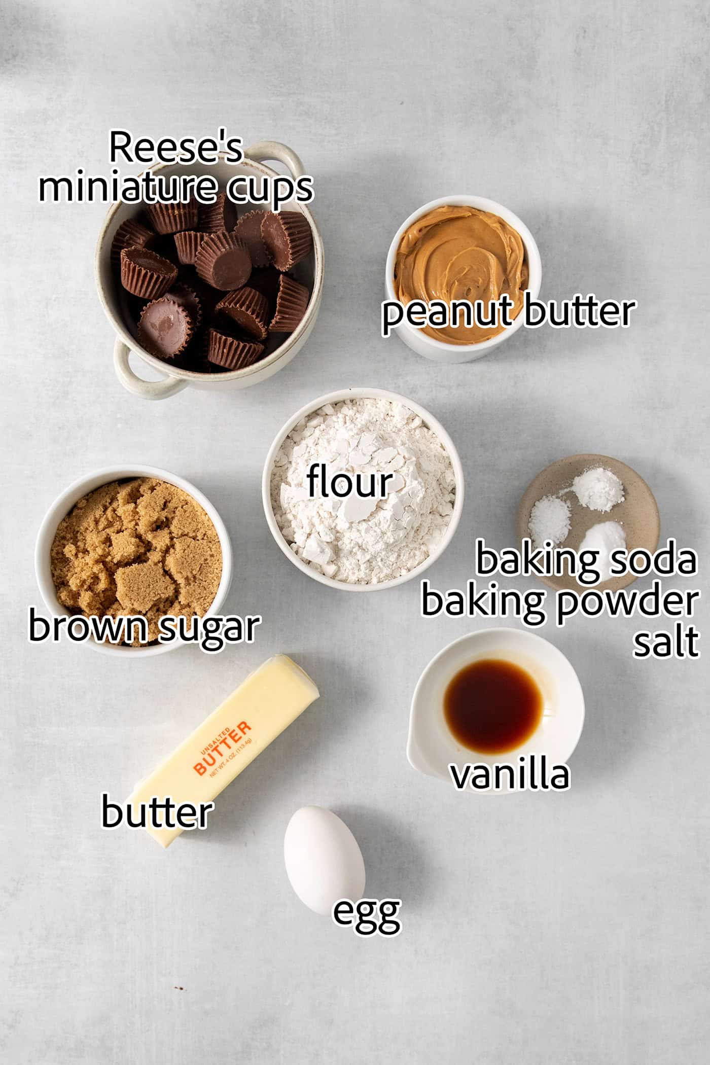 Ingredients to make peanut butter cup cookies are shown labelled and portioned out: flour, vanilla, peanut butter, egg, butter, baking powder, baking soda, salt, brown sugar, mini peanut butter cups.