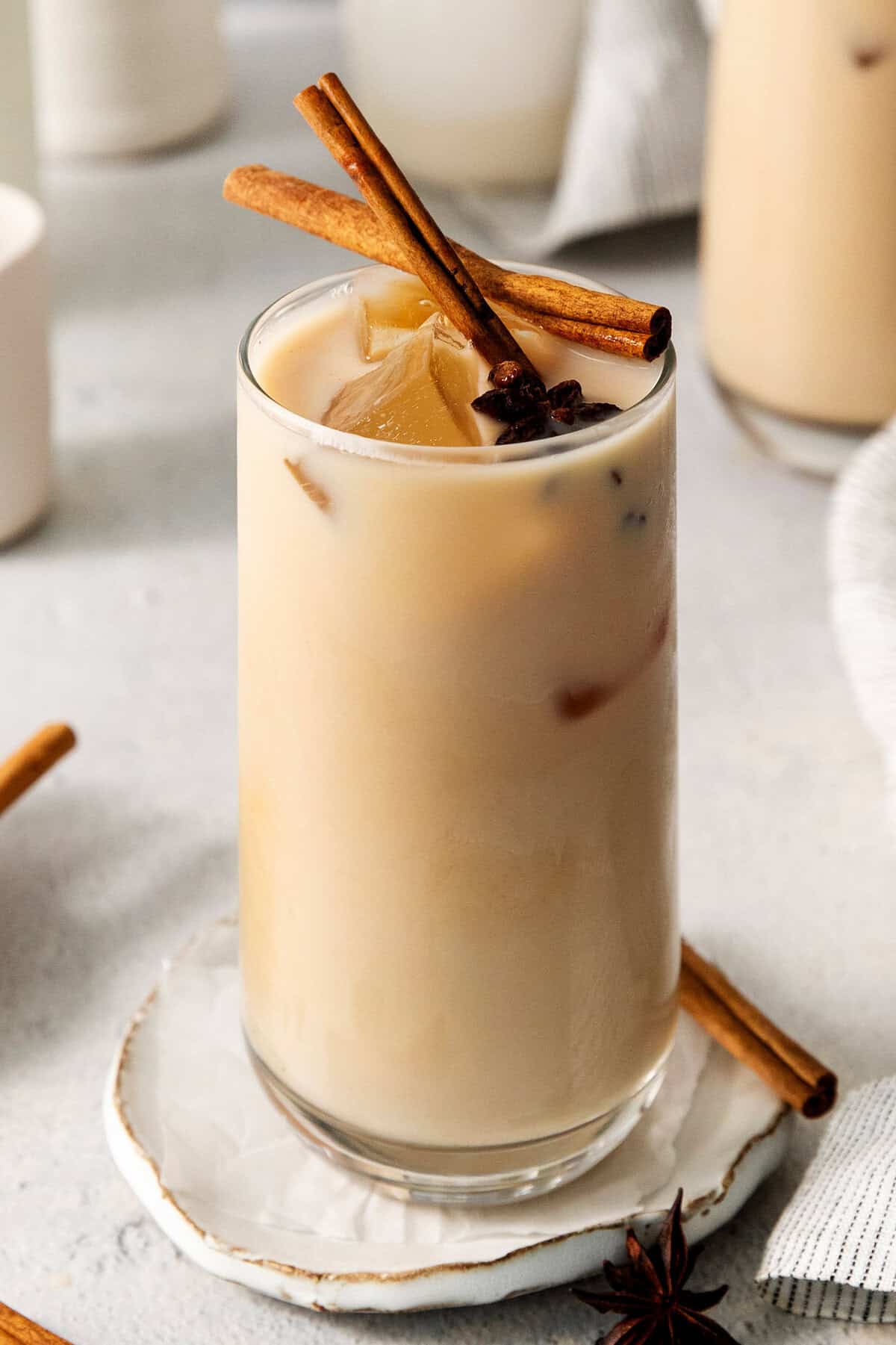 This easy Iced Chai Latte recipe is the perfect refreshing drink, featuring crisp black tea, warm spices, and a hint of brown sugar. A tall, milky glass of iced chai latte is served on a white plate and garnished with cinnamon sticks and star anise.