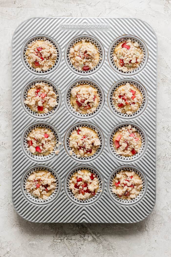 a muffin pan filled with batter that is topped with diced rhubarb and crumb topping