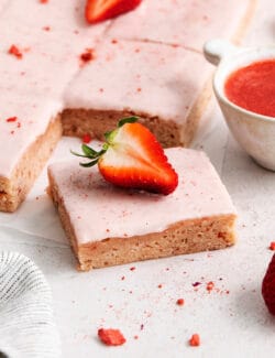 Pink icing-topped strawberry brownies are cut into squares, with strawberries scattered around them.