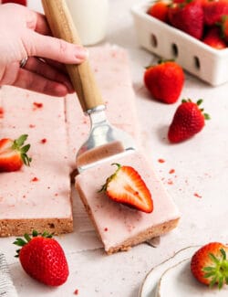 A serving spatula displays a strawberry-topped strawberry bar with pink icing.