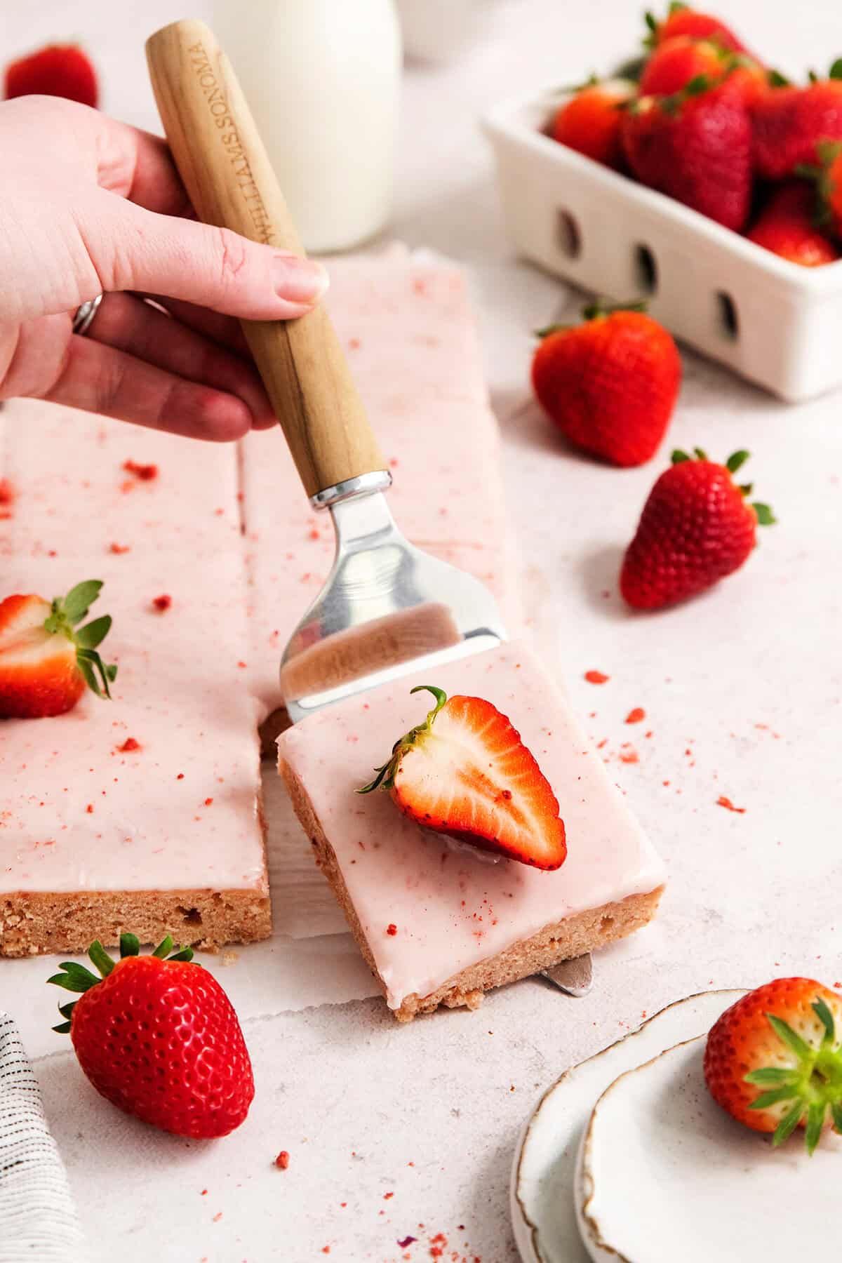 A serving spatula displays a strawberry-topped strawberry bar with pink icing.
