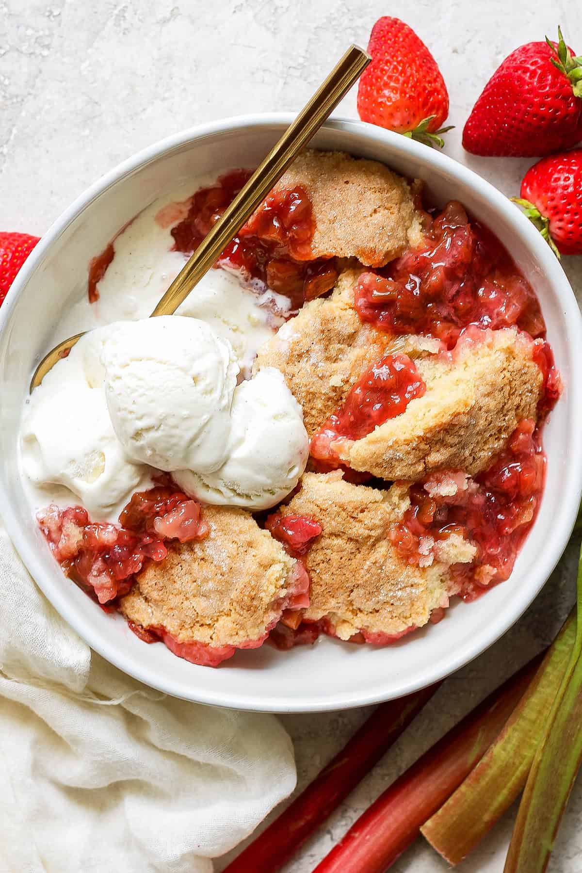 individual serving of strawberry rhubarb cobbler in a white bowl, with scoops of vanilla ice cream