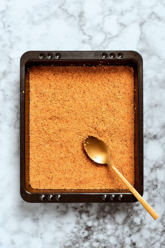 A spoon presses the graham cracker crust into a pan.