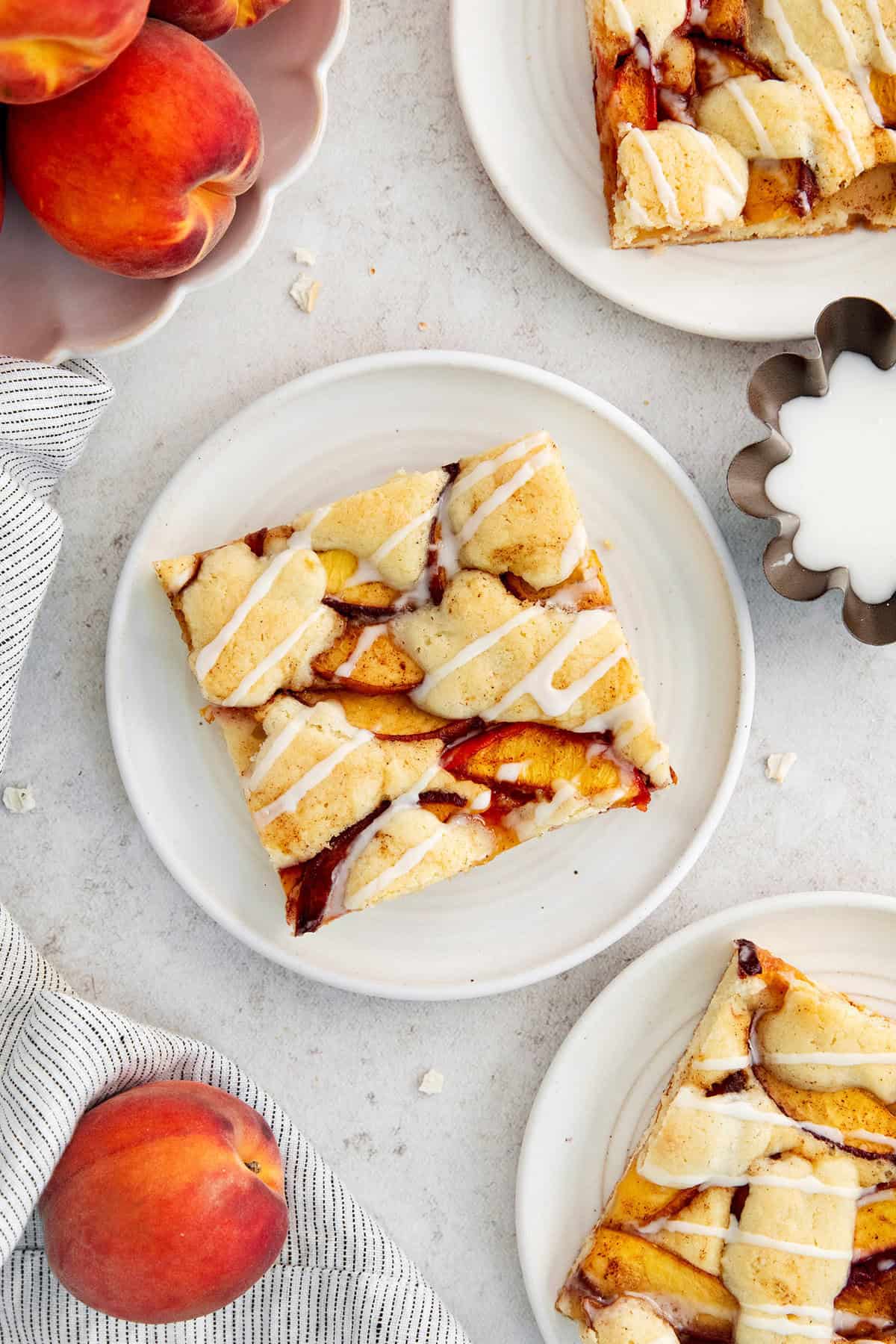 These Peach Bars feature a tender, buttery base topped with ripe, juicy peaches swirled with a touch of cinnamon. Peach bars are served on plates surrounded with whole peaches.
