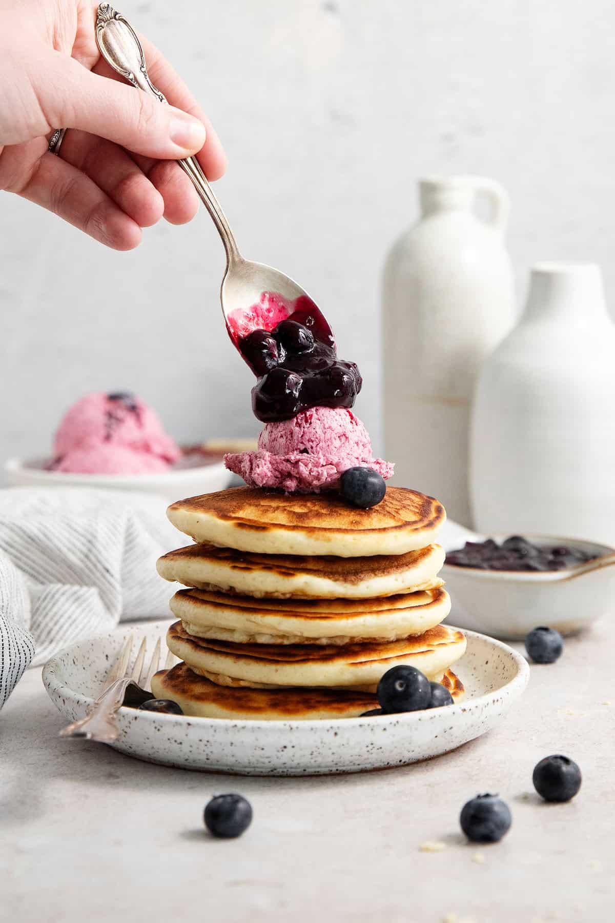 A hand spoons blueberries on top of a scoop of blueberry butter on top of a stack of pancakes.