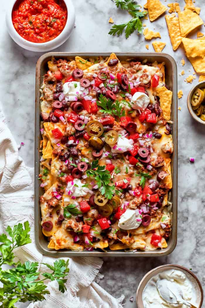 A baked tray of macho nachos surrounded with bowls of sasa, sour cream, and more chips.