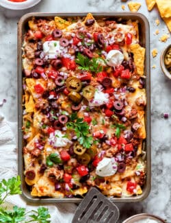 A baked tray of macho nachos topped with olives, cilantro, and sour cream.
