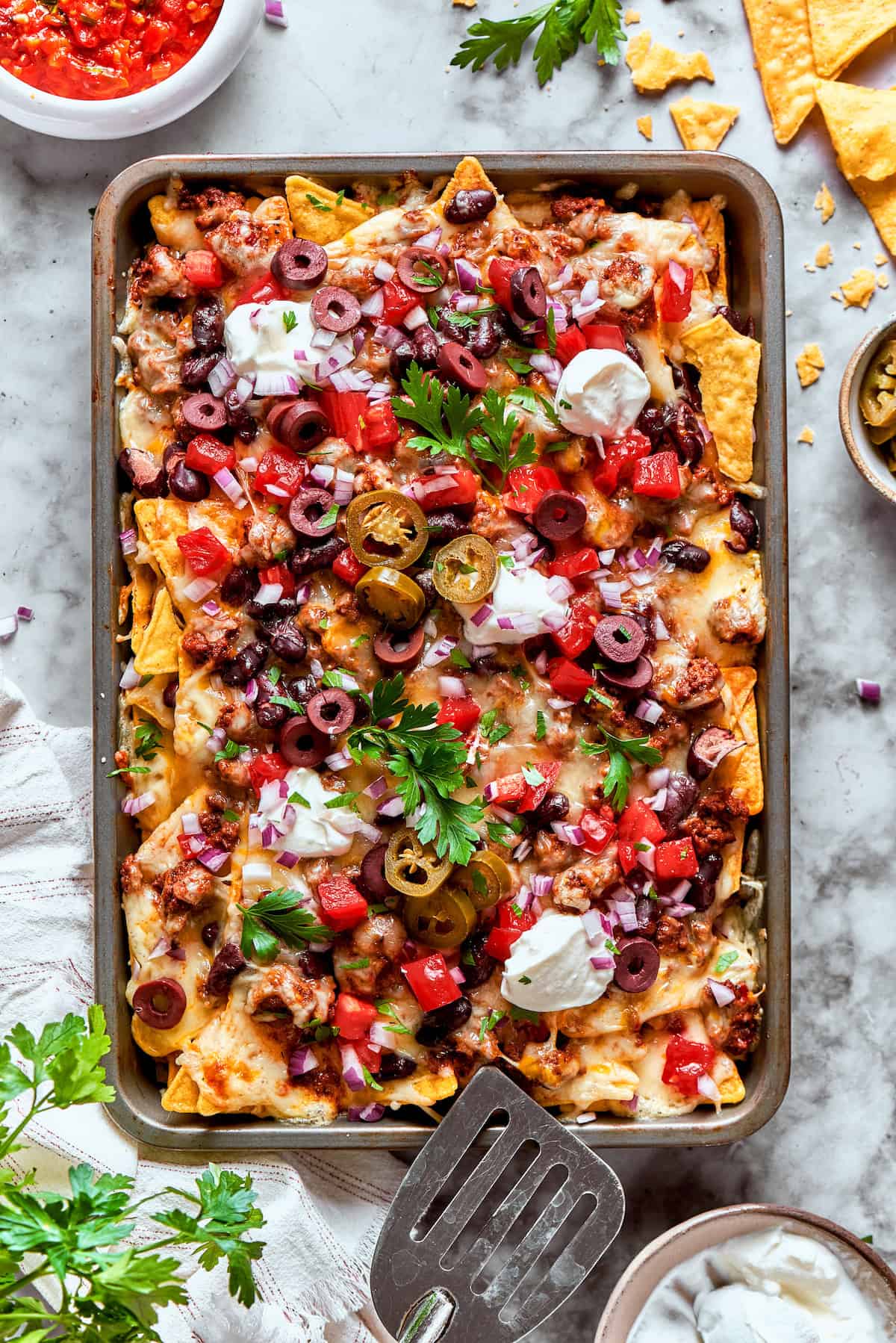 This Macho Nachos recipe features layers of crispy tortilla chips, melty cheese, and seasoned beef. Perfect for any casual gathering! A baked tray of macho nachos topped with olives, cilantro, and sour cream.