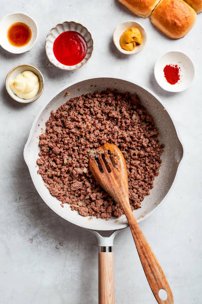 A wooden spoon stirs a pan of browned beef.