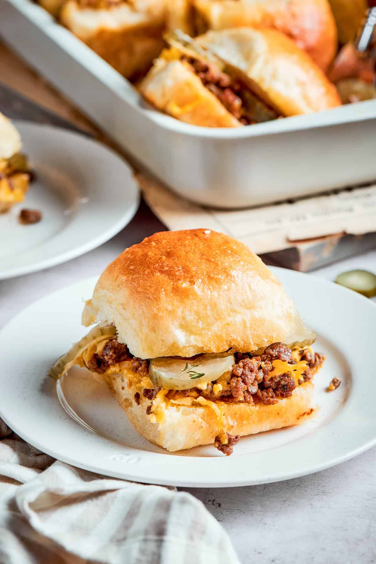 A cheeseburger slider on a white plate with a pan of sliders in the background.