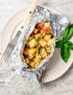Cooked potatoes peek out of a foil wrapped grilled potato packet on a plate.