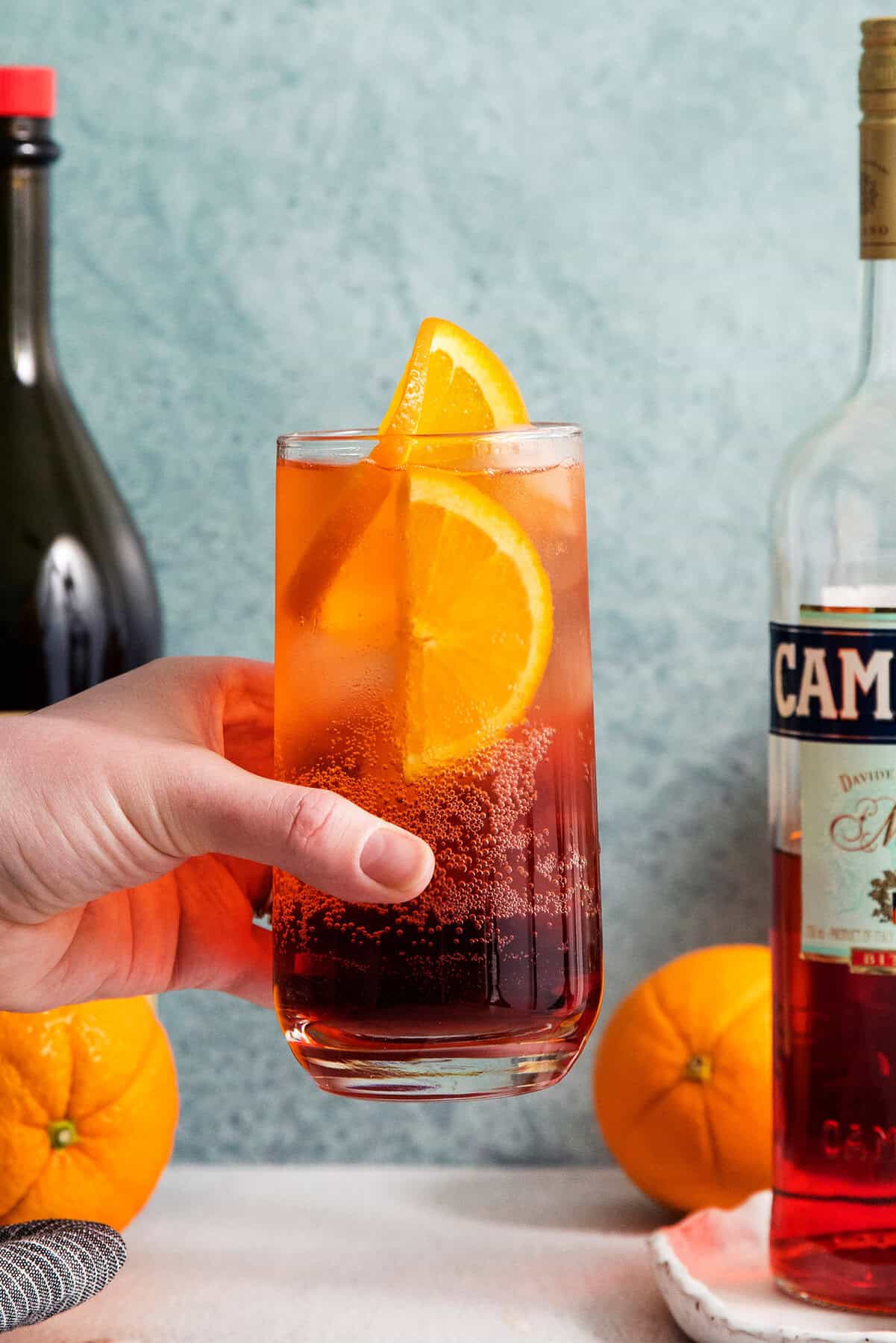 A hand holds a tall glass of Americano cocktail garnished with orange slices.