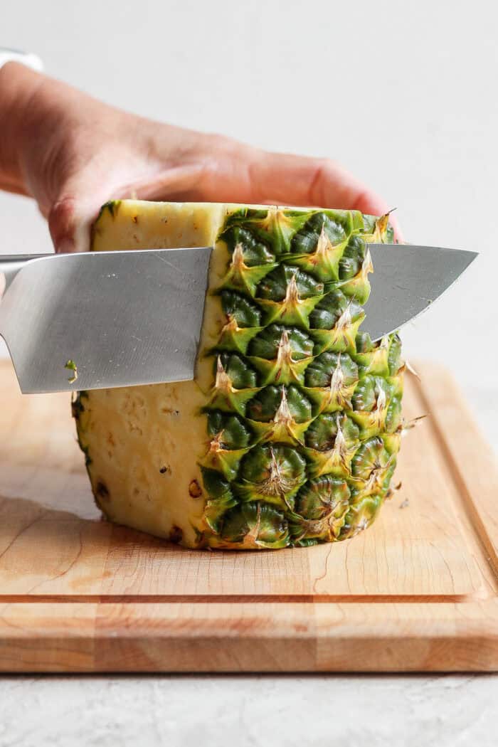 A pineapple is held upright while a knife is used to cut off the skin.