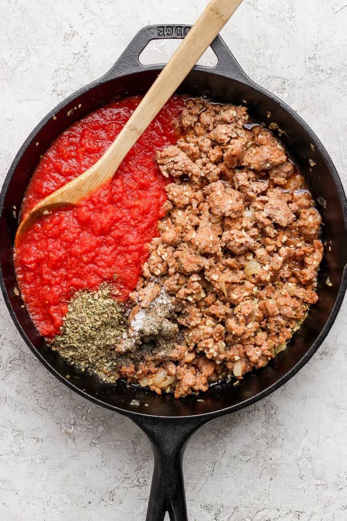 Overhead view of sausage, seasonings, and crushed tomatoes in skillet before stirring