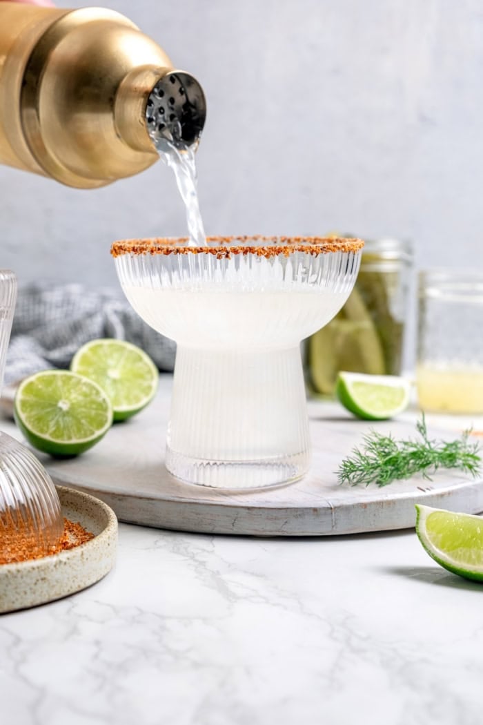 Pouring pickle margarita into glass with ice and Tajin rim