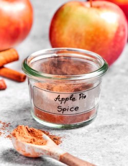 Jar of apple pie spice with spoonful of spice in foreground and apples in background
