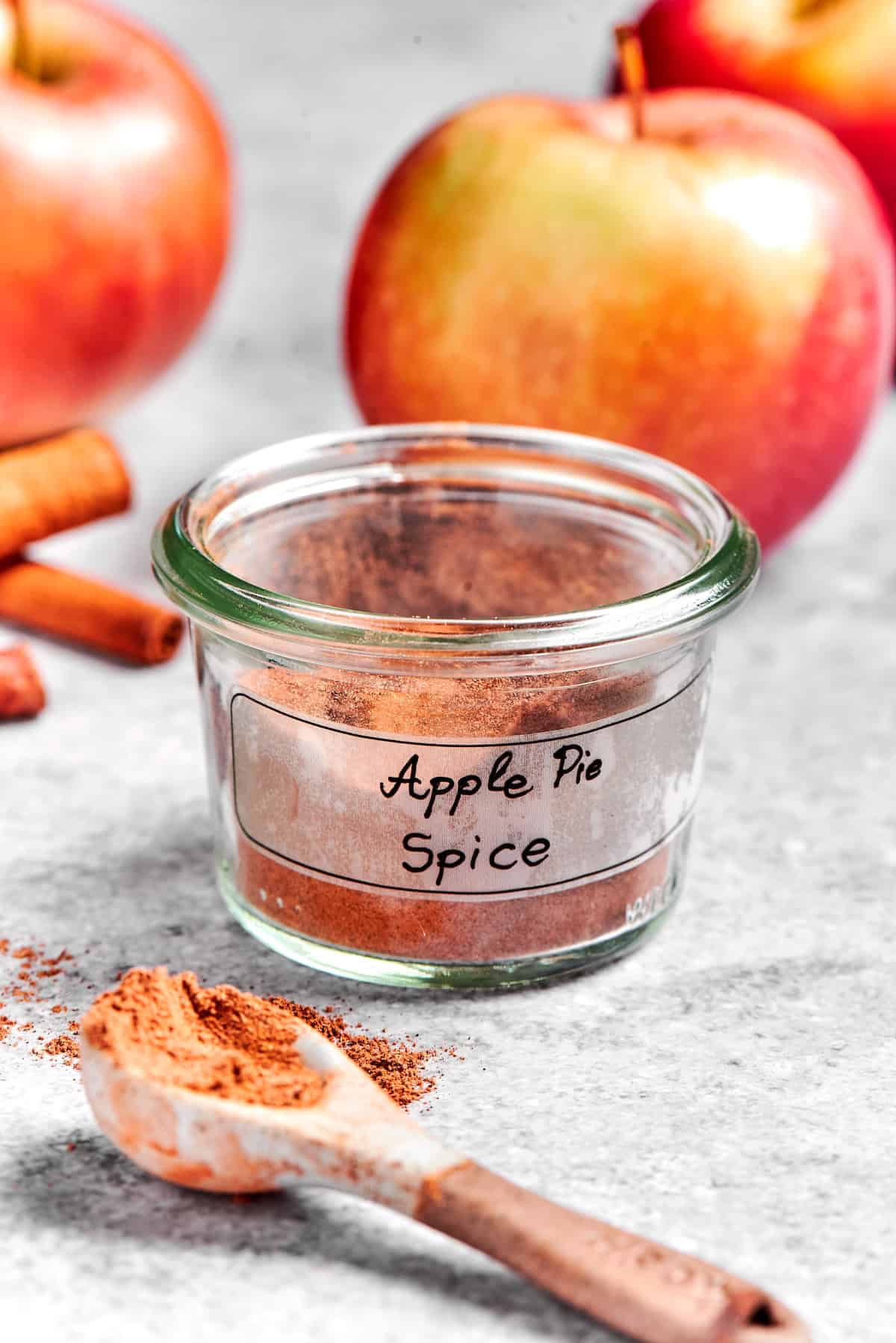 Jar of apple pie spice with spoonful of spice in foreground and apples in background