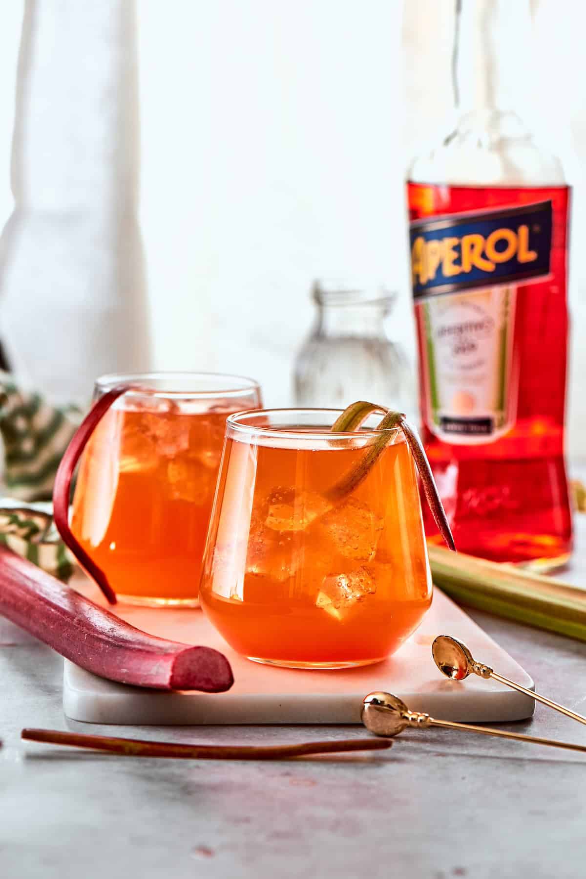 Midwest meets Italy in this summery Rhubarb Aperol Spritz! It’s a light, sparkling and refreshing cocktail perfect for using up fresh rhubarb. Two glasses of rhubarb aperol spritz cocktail with rhubarb garnish