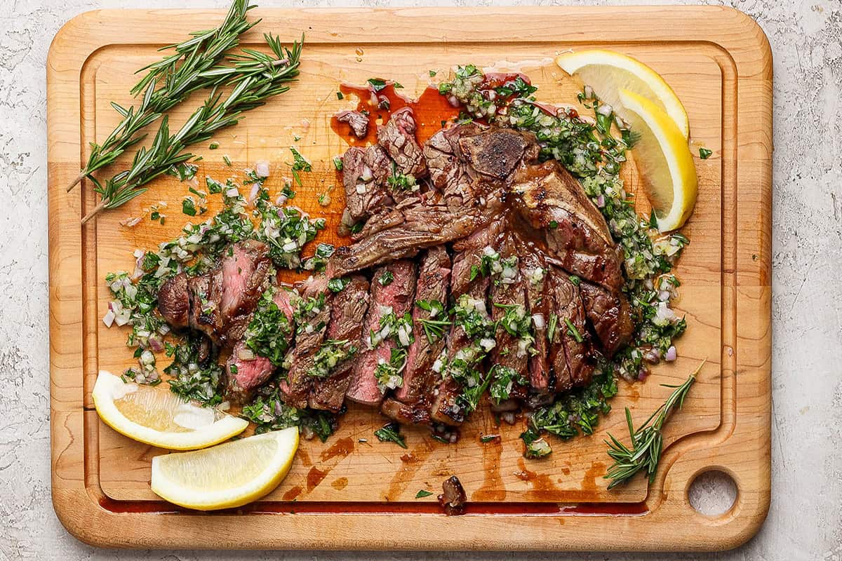 Overhead view of sliced grilled t-bone steak with board sauce, fresh rosemary, and lemon wedges