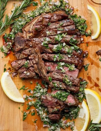 Overhead view of sliced grilled t-bone steak with board sauce, fresh rosemary, and lemon wedges