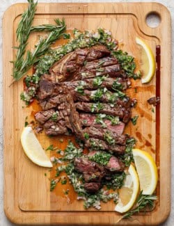 Overhead view of sliced grilled t-bone steak with board sauce, fresh rosemary, and lemon wedges