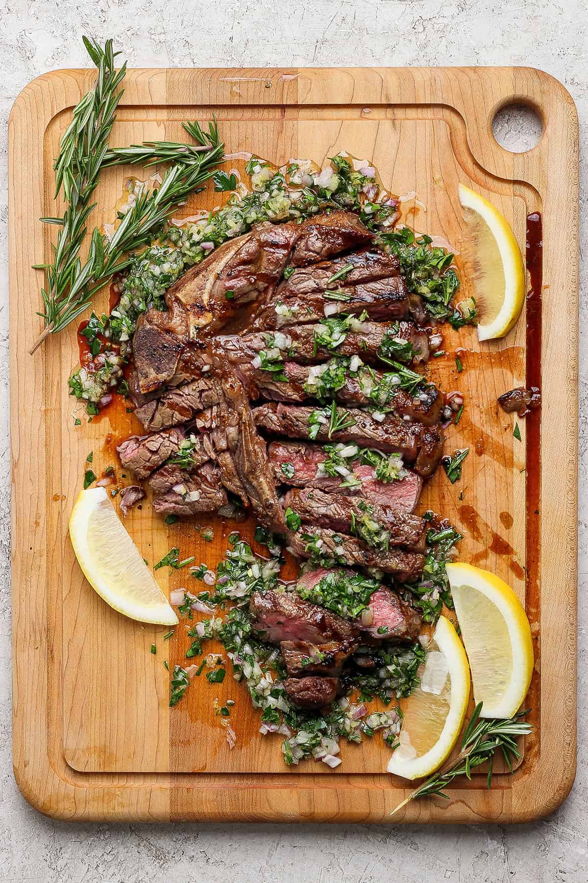 This Grilled T-Bone Steak recipe is my family's absolute favorite! Letting it soak up sauce on the cutting board makes it uniquely delicious. Overhead view of sliced grilled t-bone steak with board sauce, fresh rosemary, and lemon wedges