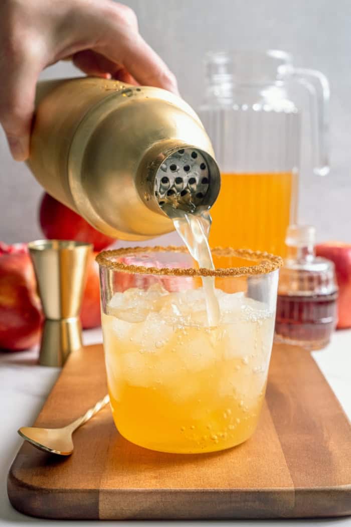 A fall spin on the classic, this Apple Cider Margarita is lightly sweetened with maple syrup and a cinnamon rim add a festive touch! Pouring apple cider margarita into glass.