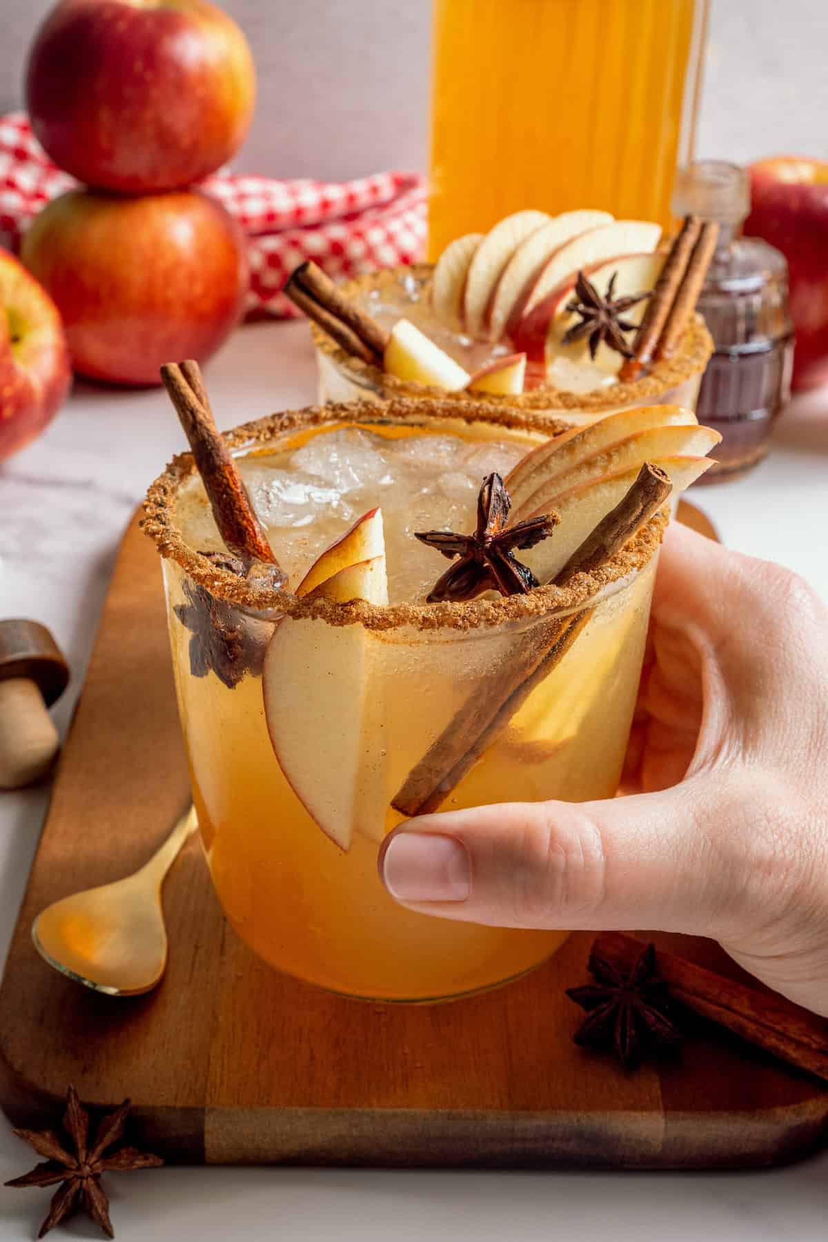 A fall spin on the classic, this Apple Cider Margarita is lightly sweetened with maple syrup and a cinnamon rim add a festive touch! Hand holding apple cider margarita in rimmed glass with apple slices, star anise, and cinnamon stick.