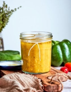 Jar of Greek salad dressing on wood board with vegetables in background