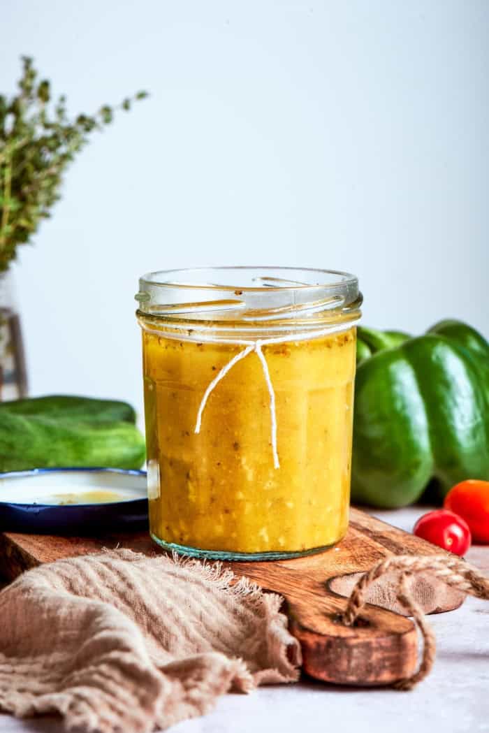 Jar of Greek salad dressing on wood board with vegetables in background