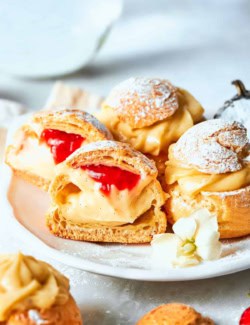 mini cream puffs filled with vanilla pastry cream on a white plate, one is cut in half to reveal strawberry sauce