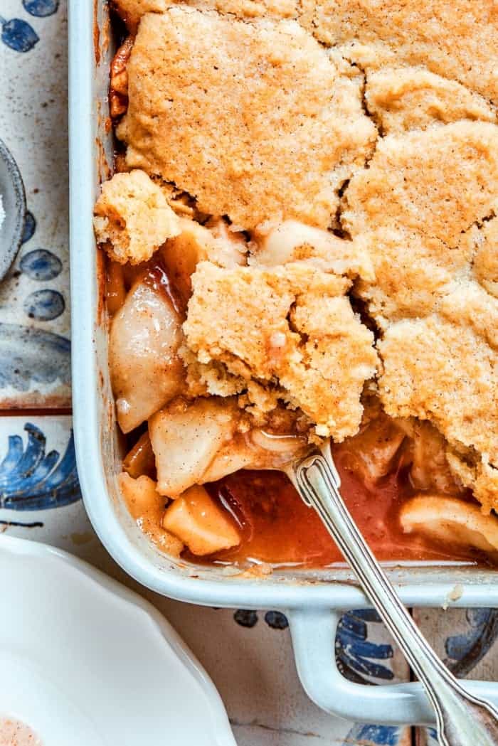 Apple Cobbler has apple pie flavor in an easier form, swapping the crust for a biscuit-like topping that bakes up tender and golden brown! a white rectangular baking dish with baked Apple Cobbler, with a spoon removing a portion