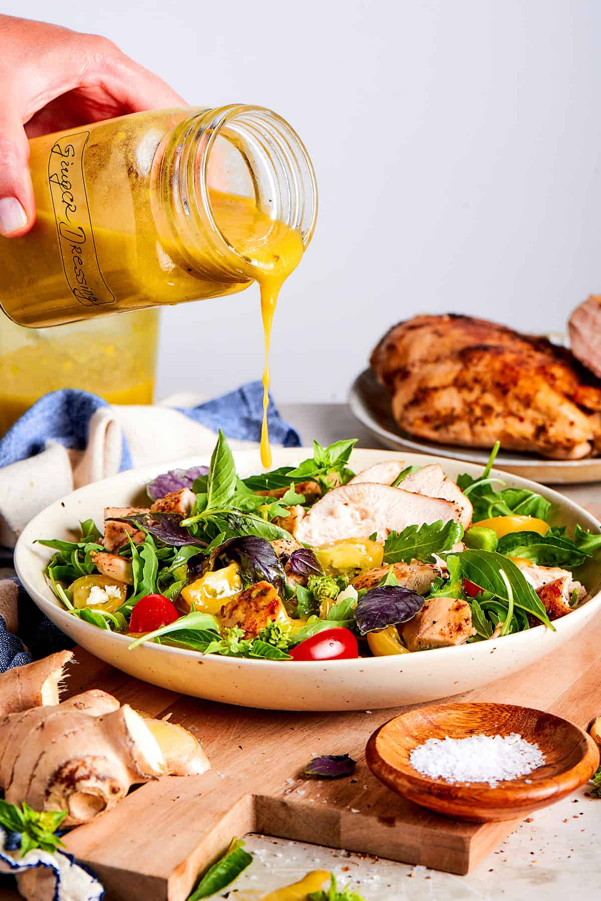 a hand pouring ginger dressing, from a jar, over a fresh green salad with grilled chicken
