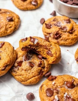 Pumpkin chocolate chip cookies on parchment paper, with one bitten to show chewy texture.
