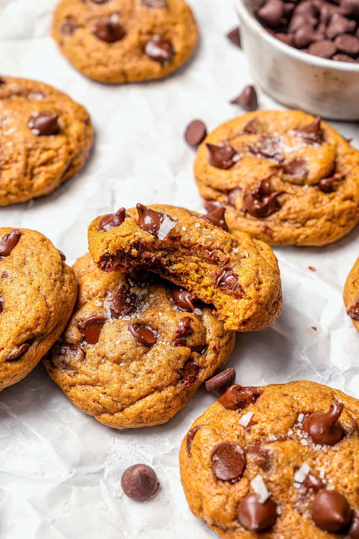 Soft, moist, and chewy, these Pumpkin Chocolate Chip Cookies are the perfect sweet treat for fall! Pumpkin pie spice adds warm, cozy flavor. Pumpkin chocolate chip cookies on parchment paper, with one bitten to show chewy texture.