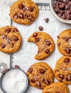 Pumpkin chocolate chip cookies on parchment lined wire rack with bowls of chocolate chips and sea salt.