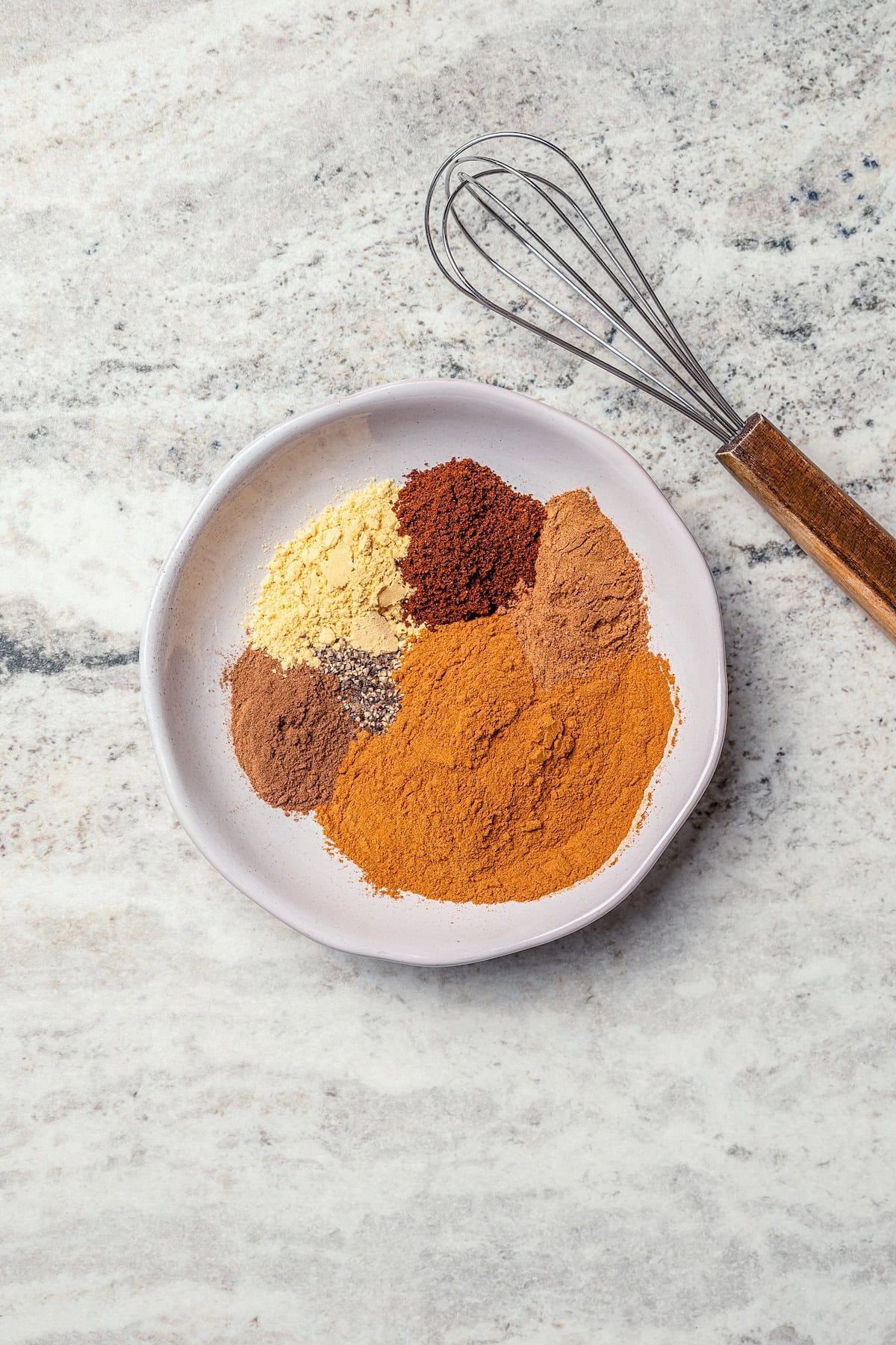 This isn't your ordinary homemade Pumpkin Pie Spice recipe! It’s rich and robust, with a touch of black pepper for some cozy warmth. Overhead view of spices for homemade pumpkin pie spice on plate with whisk.